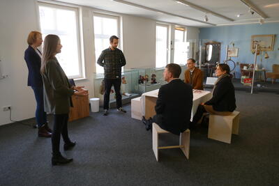 Bild vergrößern: In einem Raum sitzen drei Personen um einen Tisch herum und blicken zu drei weiteren Personen auf, die vor ihnen stehen.