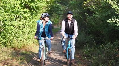 Bild vergrößern: Ein Mann und eine Frau fahren mit ihren Fahrrdern einen Waldweg entlang. Die Frau blickt den Mann lchelnd an. Beide tragen Fahrradhelme.