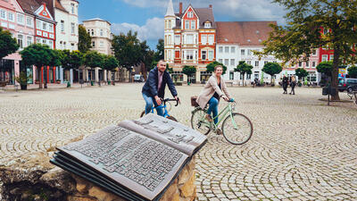 Bild vergrößern: Ein Mann und eine Frau fahren auf ihren Rdern ber das Kopfsteinpflaster einer Stadt. Im Hintergrund sind Huser zu sehen, im Vordergrund ein Bronzebuch.