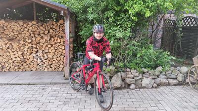 Bild vergrößern: Ein junger Mann mit Fahrradhelm steht neben einem roten Fahrrad und stützt sich mit beiden Unterarmen auf dem Lenker auf. Er blickt lächelnd in die Kamera. Im Hintergrund ist gestapeltes Brennholz und Bepflanzung zu sehen.