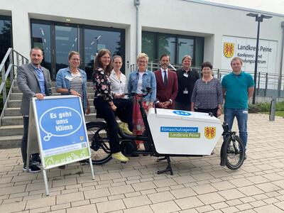 Bild vergrößern: Fnf Frauen und vier Mnnder stehen versetzt zueinander vor einem Gebude und lcheln in die Kamera. Im Vordergrund sind ein Lastenfahrrad sowie eine Aufsteller mit einer Veranstaltungsankndigung zu sehen.