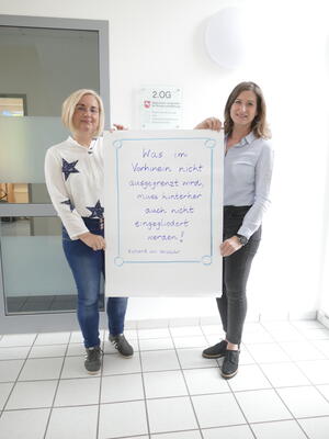 Bild vergrößern: Zwei Frauen halten gemeinsam ein groes, weies Plakat mit handschriftlichen Bemerkungen hoch.