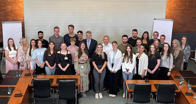 Bild vergrößern: Eine groe Gruppe von Frauen und Mnnern steht versetzt zueinander in einem Sitzungssaal vor einer weien Leinwand. Rechts und linke von der Leinwand stehen Flipcharts. Im Vordergrund sind Sitzreihen zu erkennen.