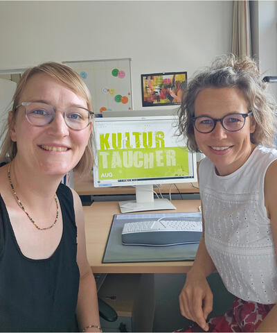 Bild vergrößern: Im Vordergrund sitzen zwei Frauen an einem Schreibtisch und wenden sich lchelnd der Kamera zu. Auf dem Schreibtisch im Hintergrund steht ein Monitor, auf dem das "Kulturtaucher"-Logo zu sehen ist.