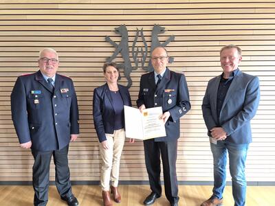 Bild vergrößern: Das Foto zeigt drei Mnner und eine Frau, die vor einer Holzwand mit dem Wappen des Landkreises Peine stehen und lchelnd in die Kamera blicken. Zwei der Mnner tragen eine Feuerwehruniform, einer davon hlt eine Urkunde in den Hnden.
