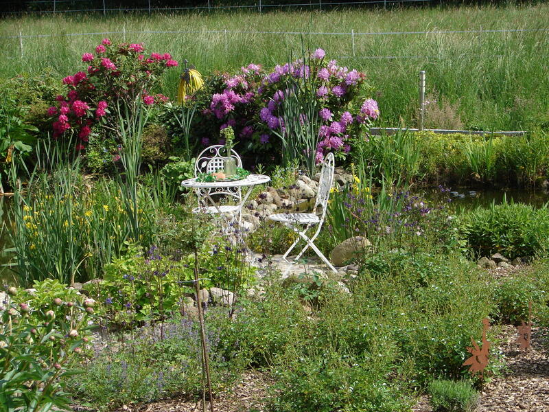 Ein weißer Metalltisch und ein weißer Metallstuhl stehen in der Mitte von einem blühenden Garten auf einem kleinen gepflasterten Bereich.