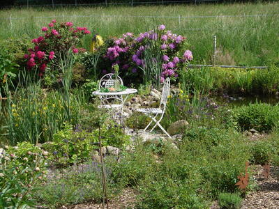 Bild vergrößern: Ein weier Metalltisch und ein weier Metallstuhl stehen in der Mitte von einem blhenden Garten auf einem kleinen gepflasterten Bereich.