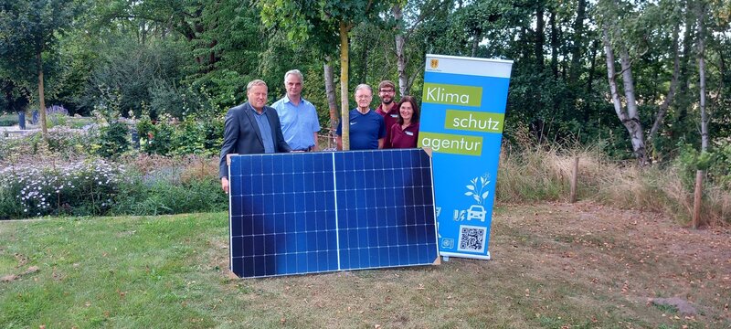 Vier Männer und eine Frau stehen nebeneinander auf einer Wiese mit Bäumen und Sträuchern im Hintergrund. Vor sich stehen die Solarpaneele eines Balkonkraftwerks, neben der Grußße steht ein Aufsteller der Klimaschutzagentur des Landkreises Peine.