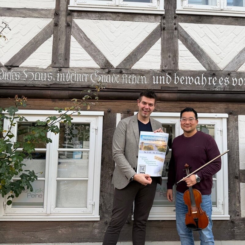 Zwei Männer stehen vor einer Fachwerkfassade. Der eine Mann hält ein Plakat in den Händen, der andere Mann eine Violine.