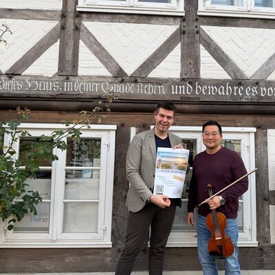 Bild vergrößern: Zwei Mnner stehen vor einer Fachwerkfassade. Der eine Mann hlt ein Plakat in den Hnden, der andere Mann eine Violine.