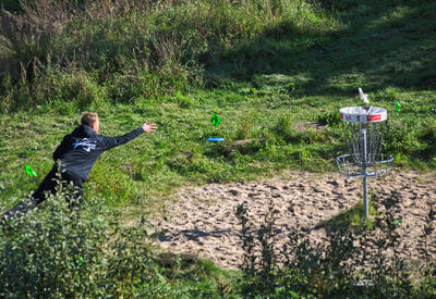 Bild vergrößern: Am linken Bildrand steht ein Mann auf einer Wiese. Er wirft eine blaue Scheibe Richtung eines Korbes auf Metall, der am rechten Bildrand in einer Sandkuhle steht. Im Vordergrund sind Bsche, im Hintegrrund hohe Grser zu sehen.