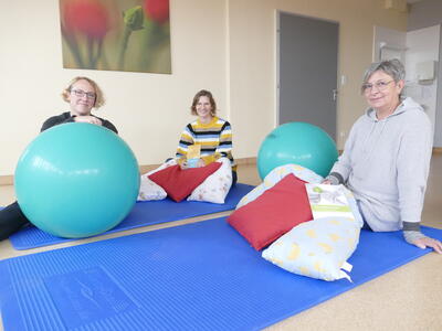 Bild vergrößern: Drei Frauen sitzen auf Turnmatten auf dem Fuboden, umgeben von Kissen und Gymnastikbllen. Alle lcheln in die Kamera.