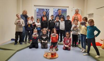 Bild vergrößern: Vier erwachsene Personen stehen im Hintergrund und blicken lchelnd in die Kamera. Im Vordergrund. Im Vordergrund sind 17 Kinder zu sehen, die teilweise stehen oder auf dem Fuboden knien. Einige der Kinder halten Bcher in die Kamera.