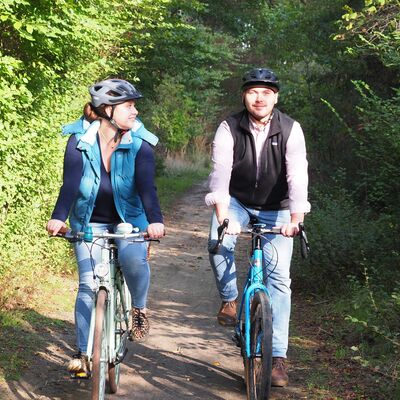 Bild vergrößern: Ein Mann und eine Frau fahren mit ihren Fahrrädern einen Waldweg entlang. Die Frau blickt den Mann lächelnd an. Beide tragen Fahrradhelme.