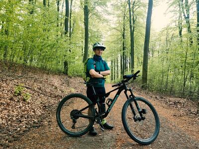 Bild vergrößern: Ein Mann steht mit verschrnkten Armen hinter seinem Mountainbike auf einem Waldweg und lchelt in die Kamera.