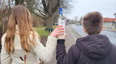 Bild vergrößern: Ein Mädchen (links) und ein Junge (rechts) stehen an einer Straße mit dem Rücken zur Kamera. Zwischen ihnen halten sie gemeinsam eine Flasche mit Alkohol.