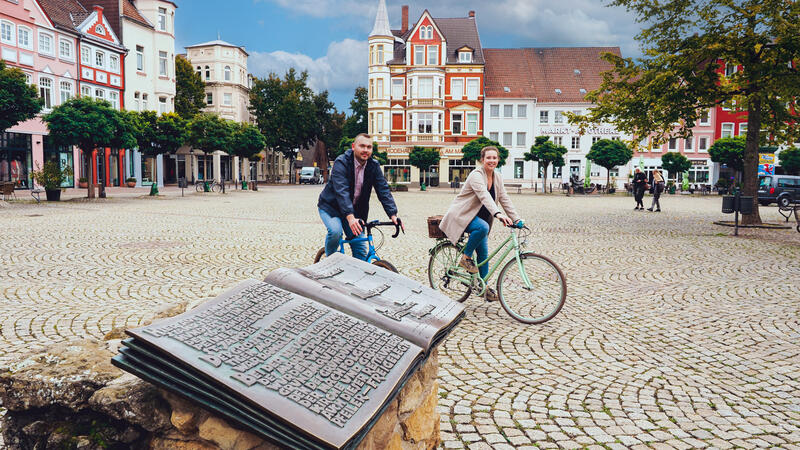Ein Mann und eine Frau fahren auf ihren Rädern über das Kopfsteinpflaster einer Stadt. Im Hintergrund sind Häuser zu sehen, im Vordergrund ein Bronzebuch.