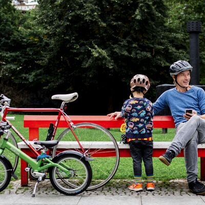 Bild vergrößern: Ein Mann mit Fahrradhelm sitzt auf einer roten Bank, der Kamera zugewandt neben ihm steht ein Kind, welches ebenfalls einen Fahrradhelm trägt. An die Bank sind ein rotes Herrenrad und ein gründes Kinderrad gelehnt. Im Hintergrund ist eine Wiese und ein Wald zu sehen.
