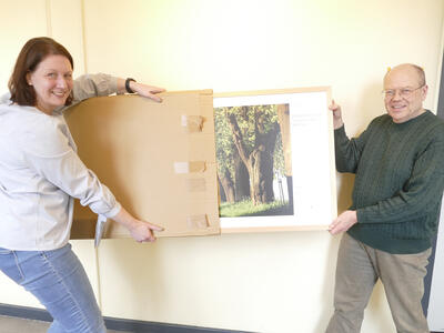 Bild vergrößern: Eine Frau und ein Mann stehen vor einer Wand und entpacken gemeinsam ein groformatiges Foto aus einem Karton.