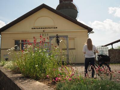 Bild vergrößern: Eine Frau steht mit dem Rcken zur Kamera neben einem Fahrrad. Im Hintergrund ist ein gelbes Gebude zu erkennen. Vorne links im Bild sieht man weie und pinke Blumen.
