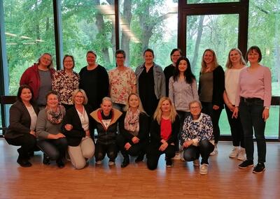 Bild vergrößern: 17 Frauen stehen in einer Gruppe vor einem groen Fenster und blicken lchelnd in die Kamera.
