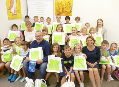 Bild vergrößern: Eine Gruppe von 22 Kindern und 4 Erwachsenen sitzen in Reihen nebeneinander und blicken lchelnd in die Kamera. Die Kinder halten weie Taschen mit grnem Aufdruck in ihren Hnden.