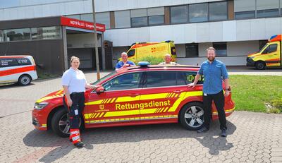 Bild vergrößern: Auf dem Bild ist ein rot-gelber Kombi mit Blaulicht auf dem Dach zu sehen. Seitlich vor dem Fahrzeug stehen ein Mann und eine Frau, hinter dem Fahrzeug stehen zwei weitere Mnner. Alle Personen blicken lchelnd in die Kamera.