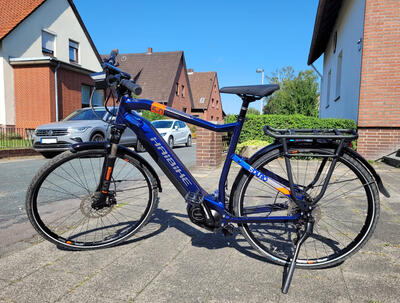 Bild vergr&ouml;&szlig;ern: Im Vordergrund ist ein blaues Fahrrad zu erkennen, welches auf einem Hof abgestellt ist. Im Hintergrund sind eine Stra�e, Autos und H�user zu sehen.