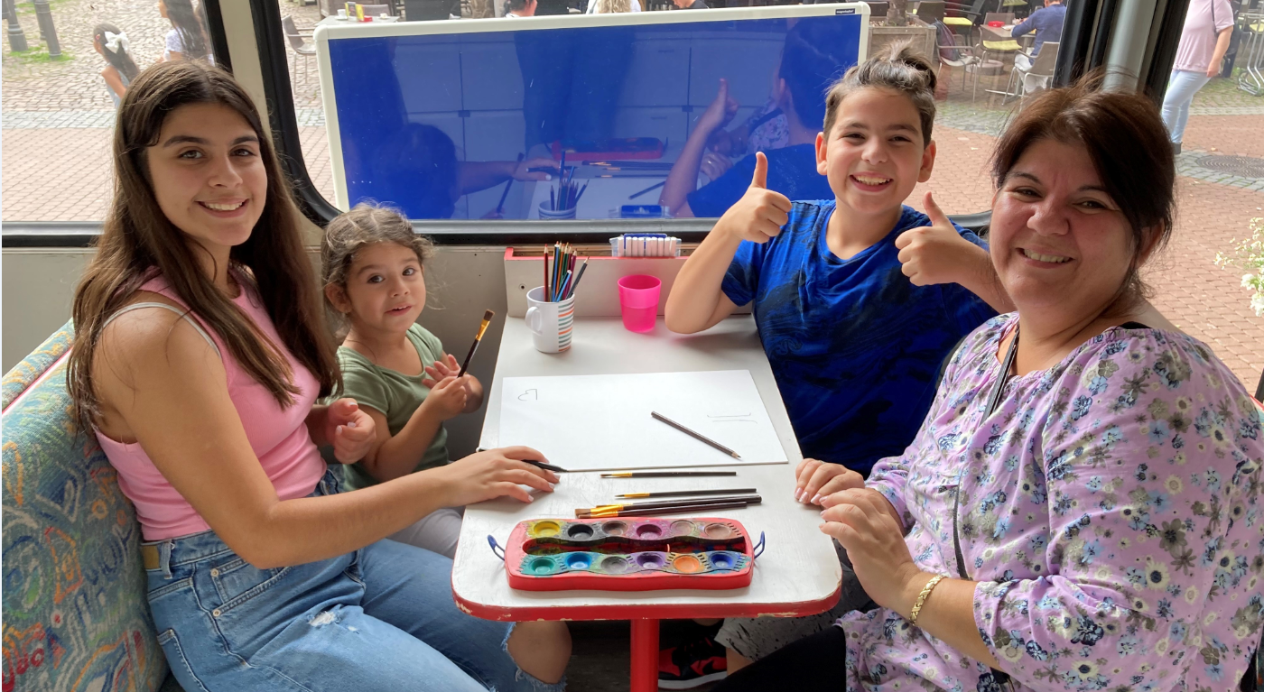 Bild vergrößern: Zwei Kinder und zwei Frauen sitzen in einem Bus an einem Tisch und lcheln in die Kamera. Auf dem Tisch liegen ein Zeichenblock und ein Tuschkasten.