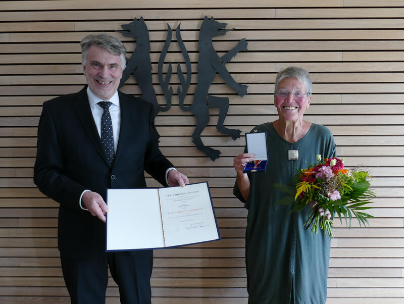 Ein Mann und eine Frau stehen vor einer vertäfelten Wand, die das Kreiswappen in schwarz zeigt. Beide lächeln in die Kamera. Der Mann hält eine Urkunde in den Händen und die Frau einen Blumenstrauß und eine geöffnete Schatulle mit einem Orden.