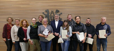 Bild vergr&ouml;&szlig;ern: 12 M�nnder und Frauen stehen vor einer holzvert�felten Wand, an der das Wappen des Landkreises Peine angebracht ist und l�cheln in die Kamera. Einige von ihnen halten Urkunden und kleine Pr�sente in ihren H�nden.