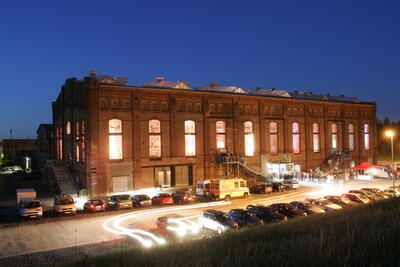 Bild vergr&ouml;&szlig;ern: Auf dem Bild ist eine erleuchtete Industriehalle vor einem blauen NAchthimmel zu sehen. Im Vodergrund sieht man parkende Fahrzeuge, auf den Eingangstreppen der Halle stehen Menschen.