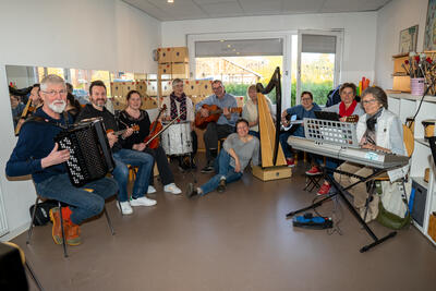 Bild vergrößern: Mehrere Musikerinnen und Musiker sitzen mit ihren Instrumenten im Halbkreis in einem hellen Raum mit weien Wnden und lcheln in die Kamera.