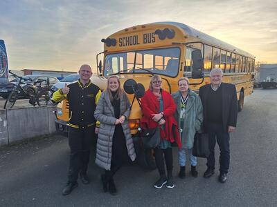 Bild vergrößern: Eine Gruppe von drei Frauen und zwei Mnnern steht vor der Motorhaube eines amerikanischen Schulbusses in gelber Lackierung. Alle lcheln in die Kamera.