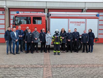 Bild vergr&ouml;&szlig;ern: Eine Gruppe von Frauen und M�nnern steht vor einem Feuerwehrauto und l�chelt in die Kamera. In der Mitte steht eine Person in Feuerwehruniform, die die neue Atemschutztechnik tr�gt.