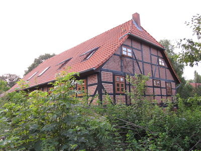 Bild vergr&ouml;&szlig;ern: Auf dem Bild ist ein Fachwerkhaus mit roten Backsteinen und rotem Satteldach zu sehen, welches von B�umen und Str�uchern umgeben ist.