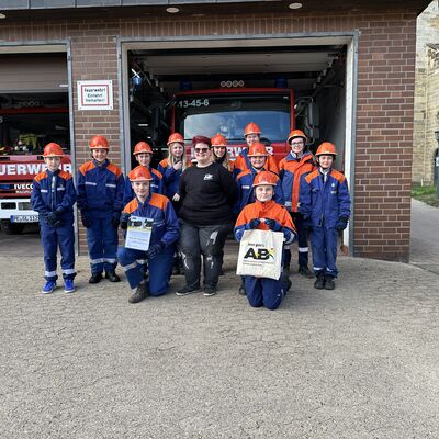 Bild vergrößern: Elf Kinder und Jugendliche in Feuerwehruniform stehen vor einer Garage mit 2 geffneten Toren in der Einsatzfahrzeuge der Feuerwehr zu sehen sind. Zwei der Kinder halten jeweils eine Stofftasche und eine Urkunde in der Hand. In der Mitte der Gruppe an Kindern steht eine junge Frau.