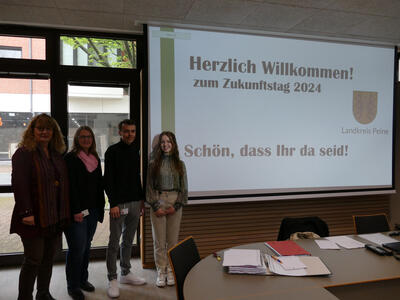 Bild vergrößern: Drei Frauen und ein Mann stehen in einem Konferenzraum neben einer Leinwand und blicken lchelnd in die Kamera.