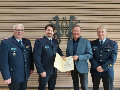 Bild vergr&ouml;&szlig;ern: Vier M�nner, drei davon in Feuerwehruniform, stehen vor einer vert�felten Wand, die das Kreiswappen in schwarz zeigt.
