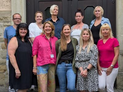 Bild vergrößern: Neun Frauen und ein Mann stehen vor einer braunen Holztr.