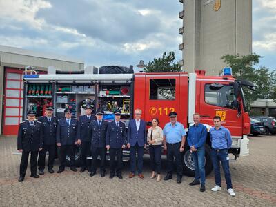Bild vergrößern: Eine Gruppe von einer Frau und zehn Mnnern, sieben davon in Feuerwehruniformen, steht vor einem Feuerwehrauto und lchelt in die Kamera.