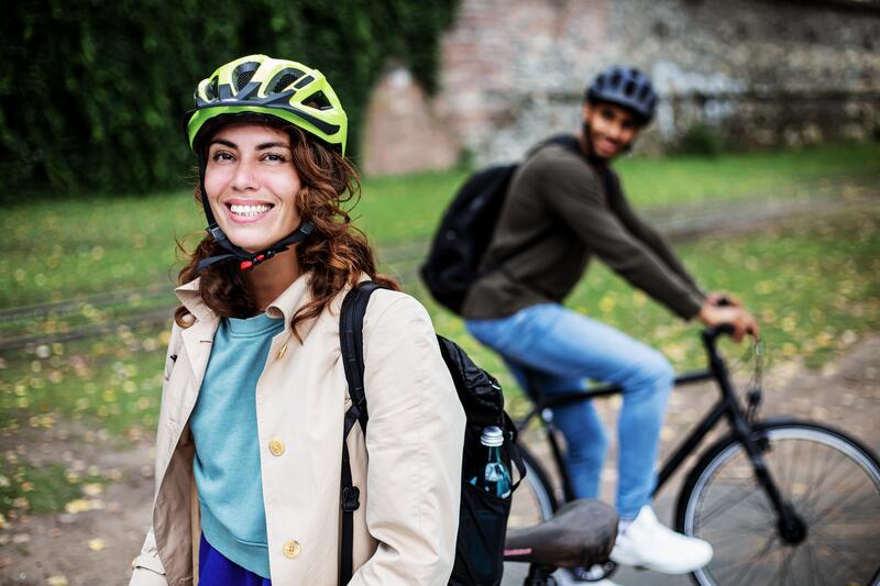 Eine Frau mit einem Fahrradhelm auf dem Kopf steht im Vordergrund und lächelt in die Kamera. Im Hintergrund ist ein Mann mit einem Fahrradhelm auf dem Kopf auf einem Fahrrad zu sehen, der die Frau lächelnd anblickt.