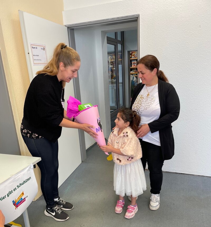 Ein Mädchen nimmt in einem Raum mit weißen Wänden eine rosa Schultüte von einer Frau entgegen. Neben dem strahlenden Mädchen steht eine weitere Frau.