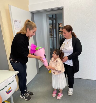 Bild vergrößern: Ein Mdchen nimmt in einem Raum mit weien Wnden eine rosa Schultte von einer Frau entgegen. Neben dem strahlenden Mdchen steht eine weitere Frau.