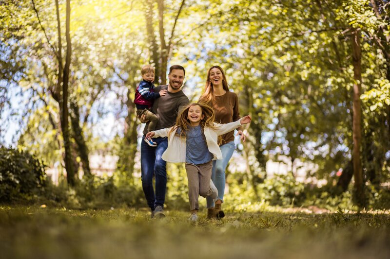 Eine Frau und ein Mann gehen mit zwei Kindern auf einem Waldweg entlang. Der Mann trägt das eine Kind auf dem Arm. Alle lachen in die Kamera. Die Sonne scheint im Hintergrund durch das Laub.