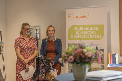 Bild vergr&ouml;&szlig;ern: Zwei Frauen stehen neben einem Stehtisch, auf dem Eine Vase mit Blumen steht. Im Hintergrund ist ein Rollup zu sehen.