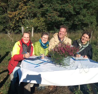 Bild vergr&ouml;&szlig;ern: Drei Frauen und ein Mann sitzen an einem Tisch mit wei�er Tischdecke und einem Heidetopf in der Natur.