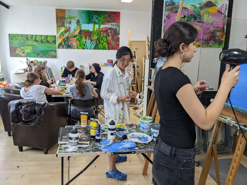 Junge Menschen sitzen in einem Atelier um einen Tisch herum. Im Vordergrund stehen zwei junge Frauen jeweils an einer Staffelei.