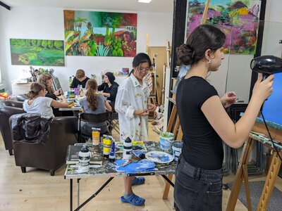 Bild vergrößern: Junge Menschen sitzen in einem Atelier um einen Tisch herum. Im Vordergrund stehen zwei junge Frauen jeweils an einer Staffelei.
