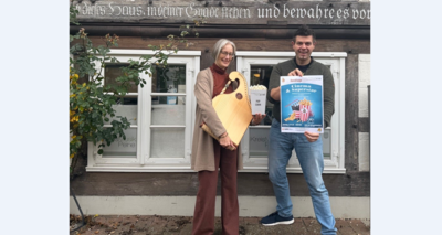 Bild vergrößern: Eine Frau und ein Mann stehen vor einer Fachwerkfassade. Die Frau hlt eine Tischharfe und eine Tte Popcorn in den Hnden. Der Mann hlt ein Plakat hoch. Beide lcheln in die Kamera.